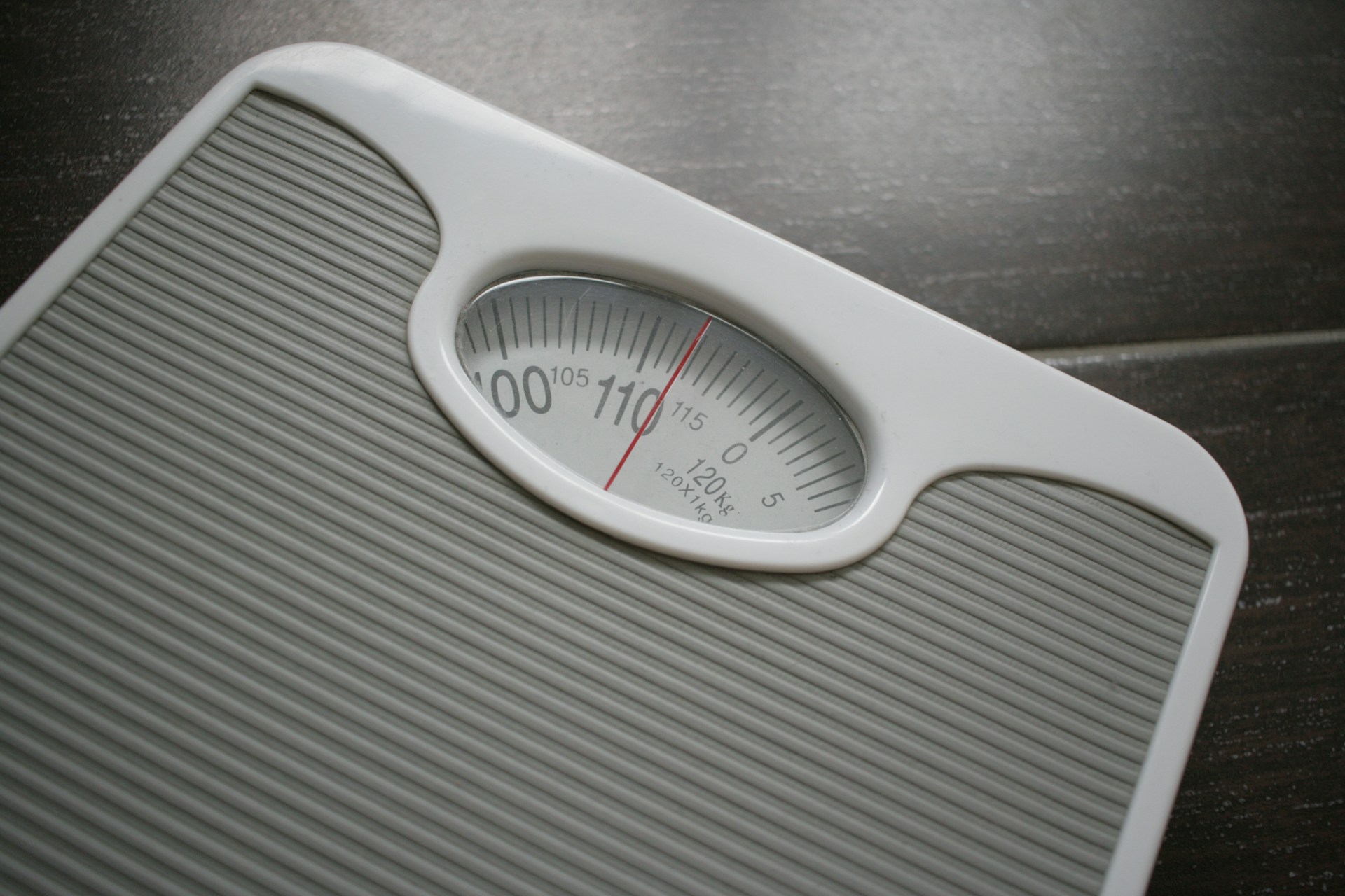 A grey analogue bathroom scale on a dark wooden floor, its needle resting on the dial.