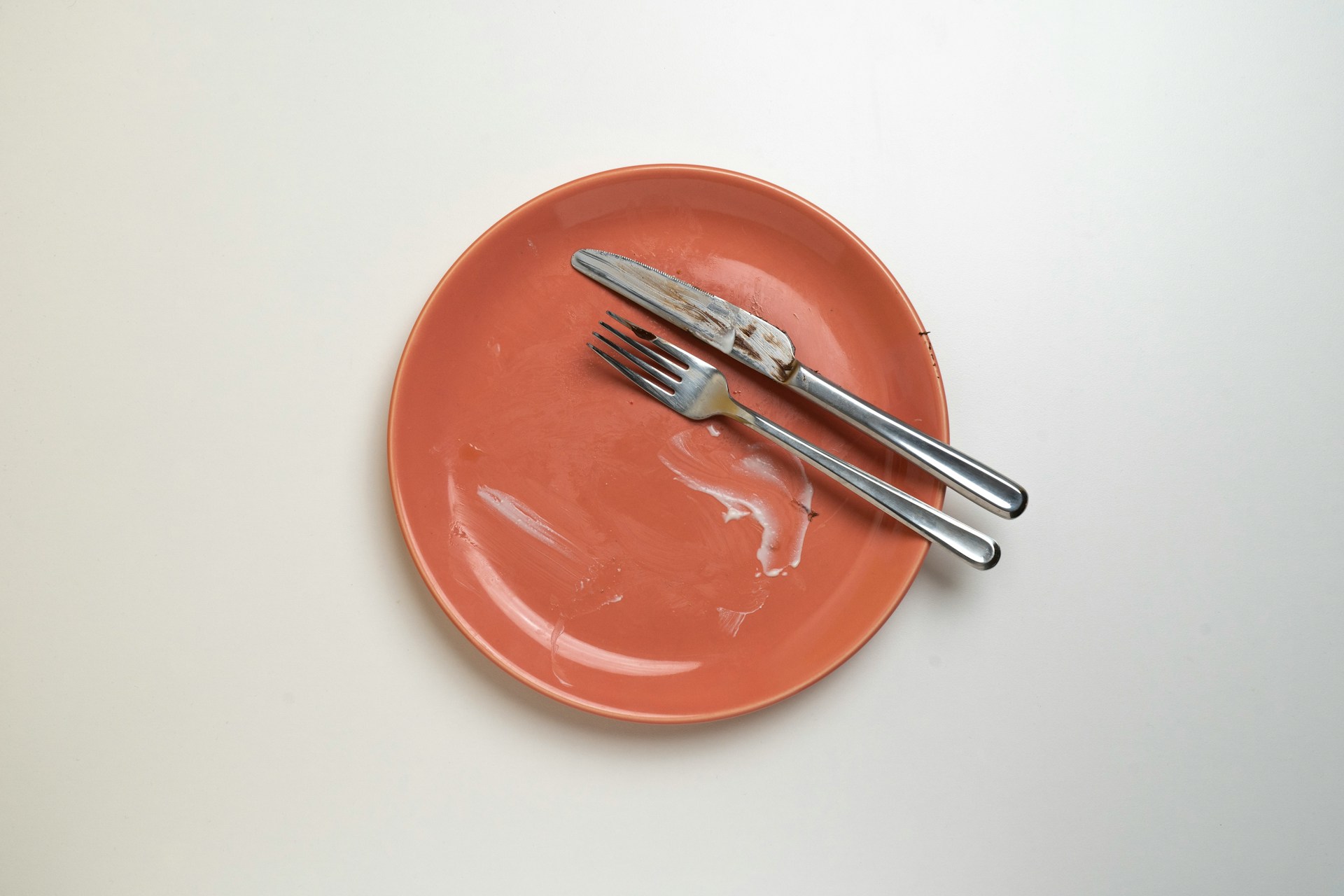 An empty pink plate with a fork and knife resting on it, on a white table.
