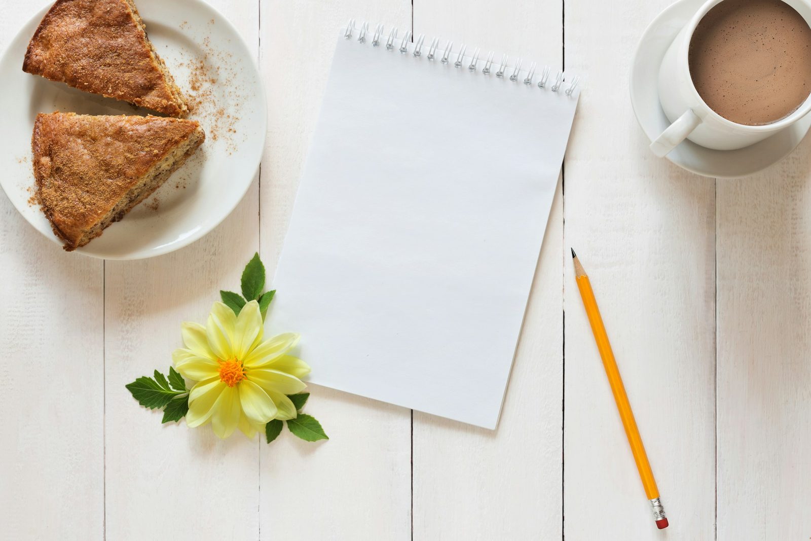 A blank spiral notepad and pencil on a white table, beside a plate of cake and a cup of cocoa.