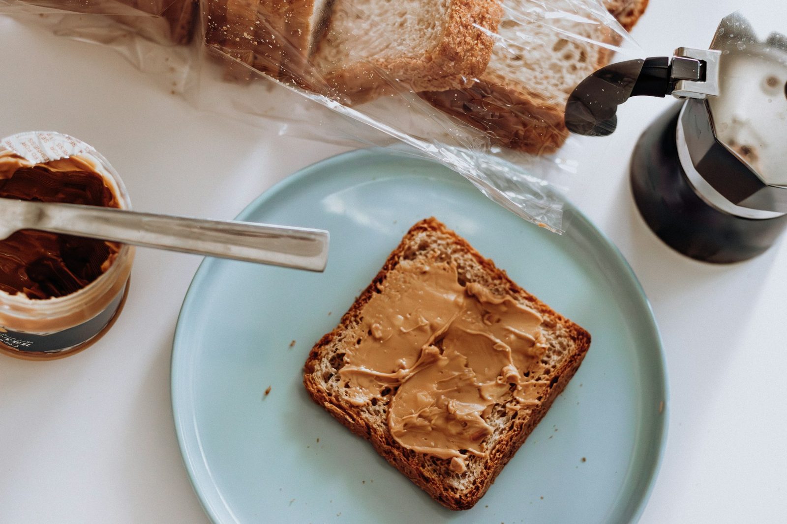 A slice of toast spread with peanut butter on a pale blue plate, next to an open jar and a coffee pot.