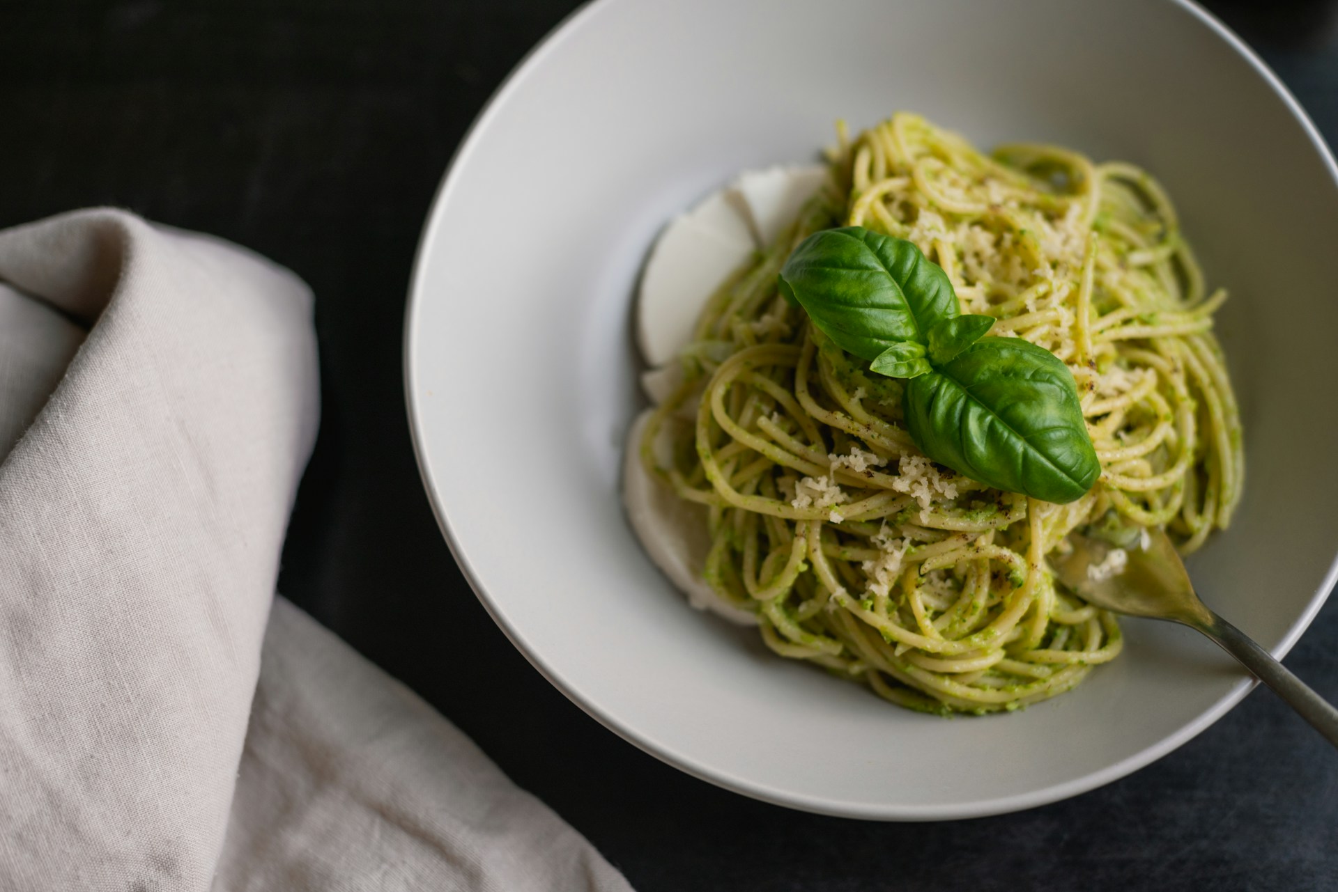 A bowl of pesto spaghetti topped with a basil leaf, next to a linen napkin on a dark table.