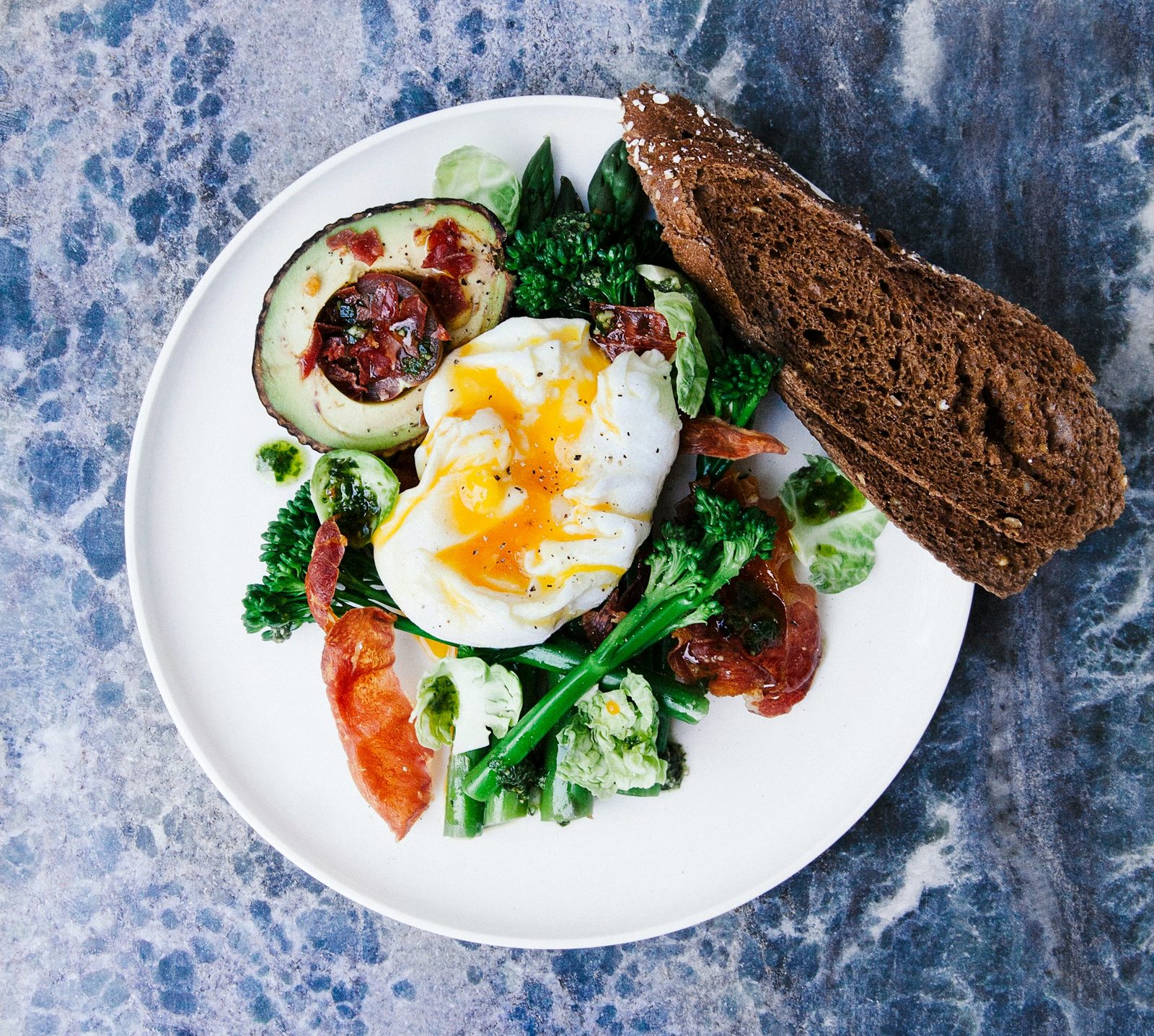 A plate with a poached egg, avocado, broccoli, tomatoes and a slice of brown bread.