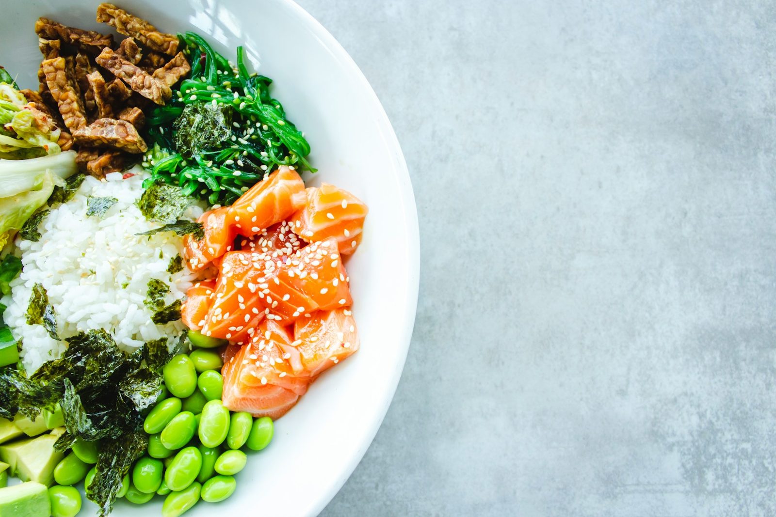 A poke bowl with salmon, rice, edamame, seaweed salad and crispy tofu on a grey table.
