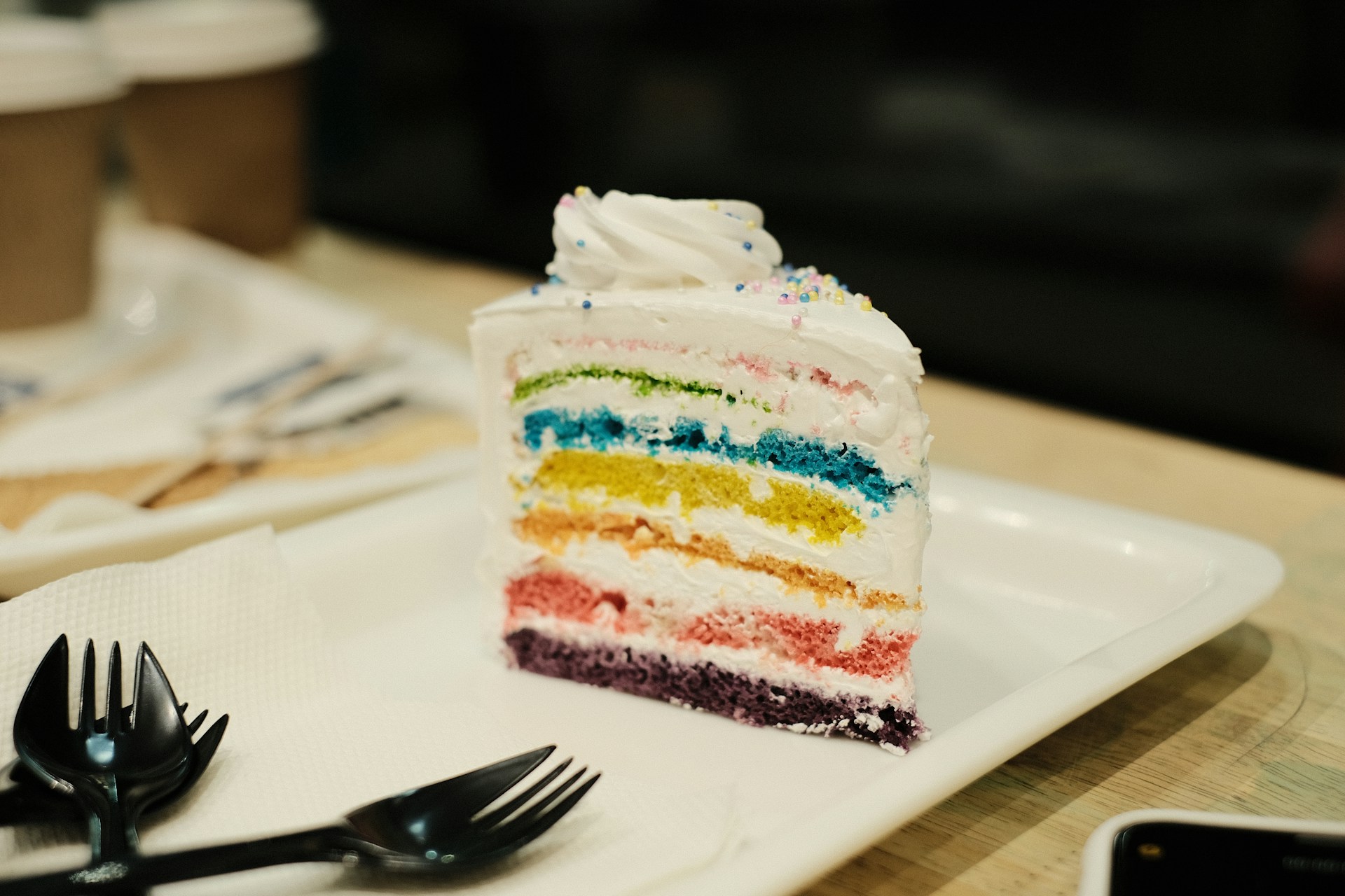 A slice of rainbow layer cake on a white plate on a cafe table.