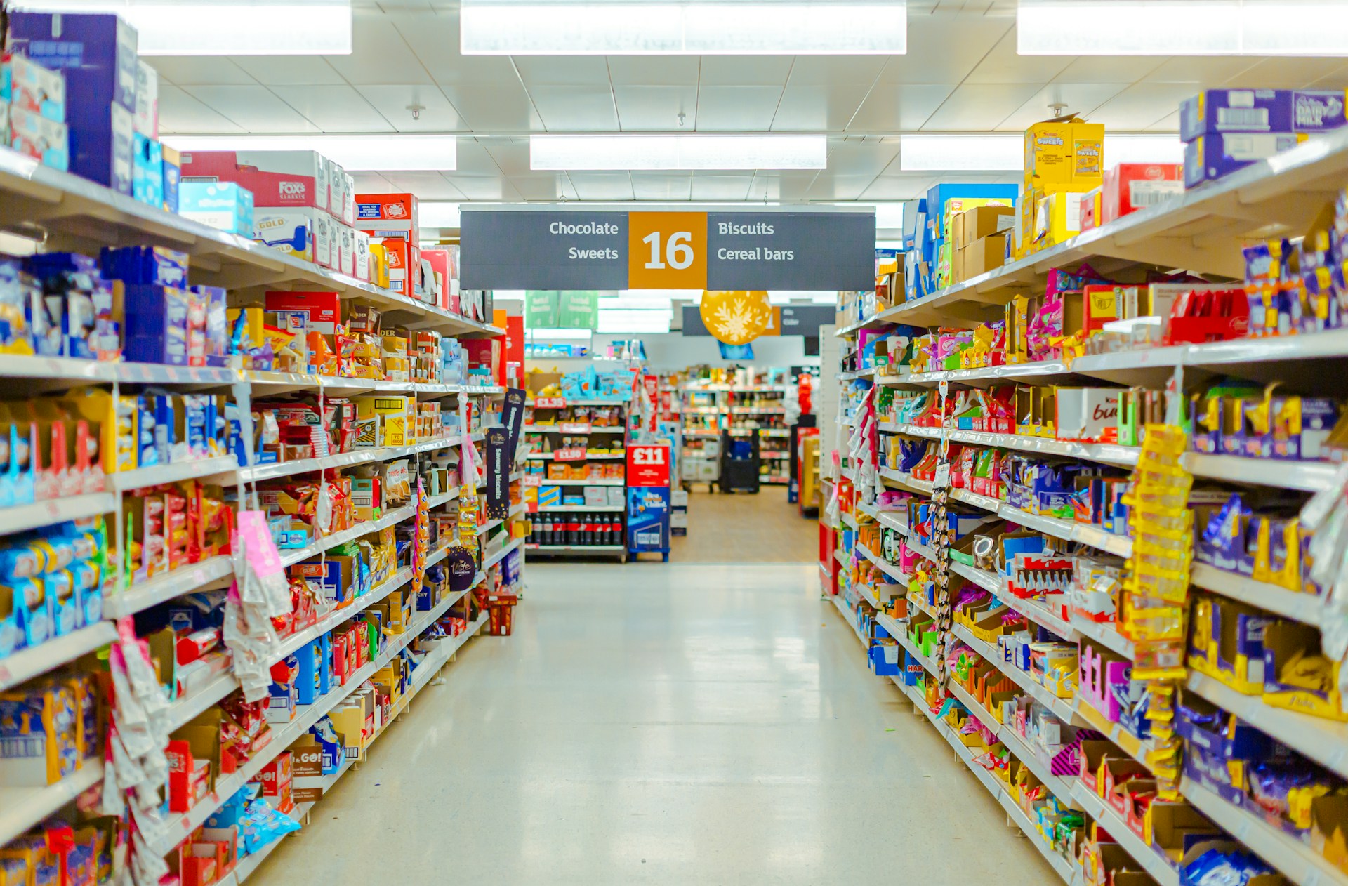 A UK supermarket aisle with shelves of biscuits, sweets and cereal bars under a sign for aisle 16.